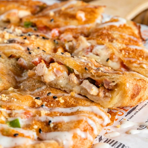 Close-up of golden-brown flatbread slices filled with chicken and vegetables, drizzled with white sauce and sesame seeds.