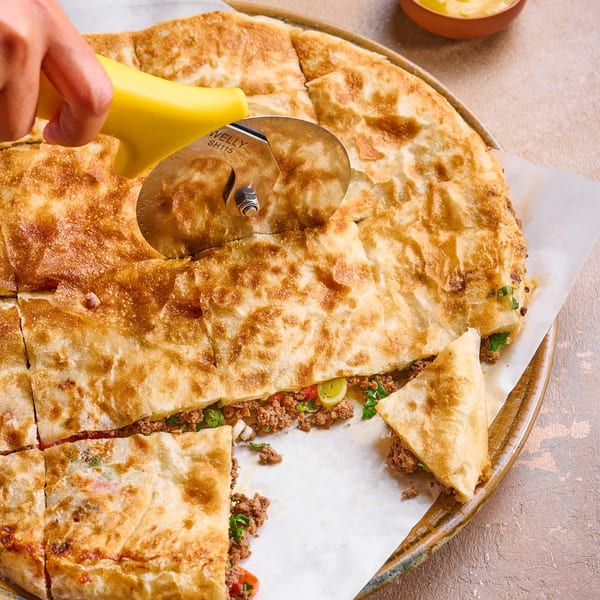 A hand uses a pizza cutter to slice a large, golden-brown savory pastry filled with ground meat.